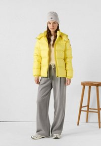 Giacca a vento gialla con cappuccio, dotata di chiusura a zip e polsini elasticizzati; indossata sopra un maglione crema e pantaloni larghi grigi. Sneakers argento.