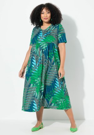 Maxikleid mit kurzen Ärmeln in Blau und Grün, mit einem blättrigen Muster. Aus weichem, leichtem Material hergestellt. Kombiniert mit grünen Ballerinas.