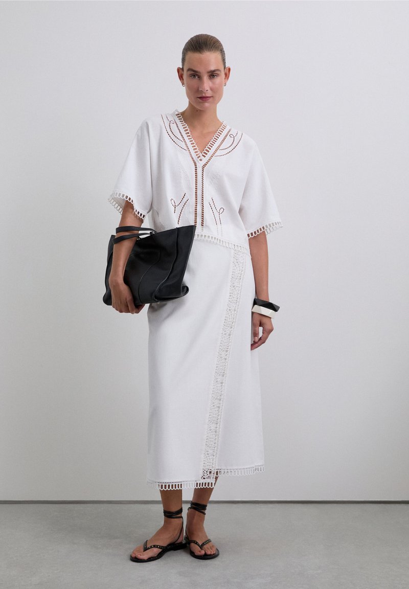Robe blanche à manches courtes, ornée de dentelle et de broderies. Associée à un sac fourre-tout en cuir noir et des sandales plates.