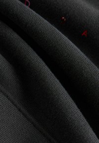 Tessuto nero con un motivo di maglia testurizzata, con strati piegati che rivelano superfici lisce e leggermente morbide. Accenti del logo multicolore visibili.