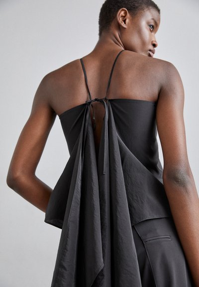 Black sleeveless top featuring a relaxed fit, back bow tie detail, and draped fabric creating a flowing effect. Smooth texture, minimal design.