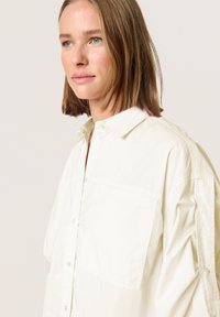Lys beige skjorte med knapper og afslappet pasform, med en enkelt brystlomme og subtil tekstur. Krave og knapper fuldender designet.