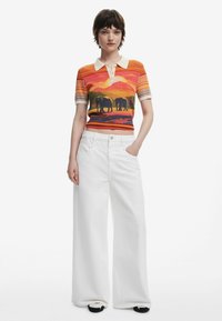 Geribbelde polo met korte mouwen in oranje, geel en grijs met een olifantenafbeelding; gecombineerd met witte wijde jeans en zwarte schoenen.