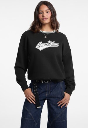 Sweatshirt noir avec le logo "Guess Jeans" blanc, col côtelé et coupe loose. Associé à un pantalon en denim foncé avec une coupe détaillée.