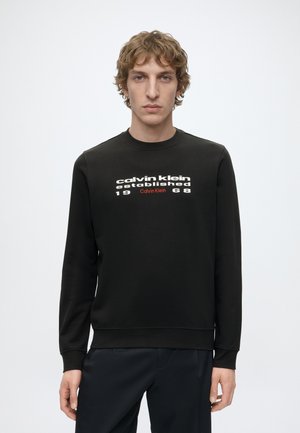 Tânăr cu păr creț care poartă un hanorac negru cu textul „calvin klein established 1968” în față, stând în fața unui fundal simplu și deschis la culoare.