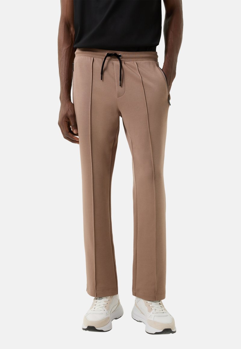 Lichtbruine, taps toelopende joggerbroek met een tailleband met trekkoord en plooidetails, gecombineerd met witte sportieve schoenen. Gladde textuur, casual ontwerp.