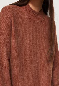 Maglione lavorato a maglia di colore ruggine con un collo alto a coste e maniche lunghe. Morbida consistenza con una vestibilità leggermente ampia, che mette in mostra un motivo a maglia grossa.