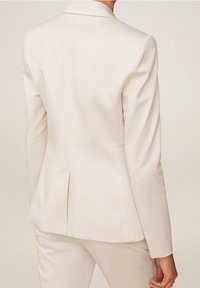 Blazer beige clair sur mesure avec une coupe structurée, des revers crantés et une fente unique à l'arrière. Texture de tissu lisse.