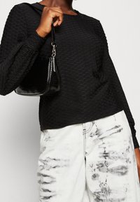 Top nero a maniche lunghe con motivo a nido d'ape, abbinato a jeans bianchi strappati con striature nere, tenuto con una piccola borsa nera.