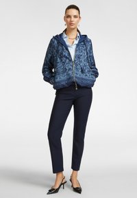 Felpa con zip blu navy con design floreale, tessuto leggero, tasche a marsupio; abbinata a pantaloni slim blu navy e scarpe col tacco nere.