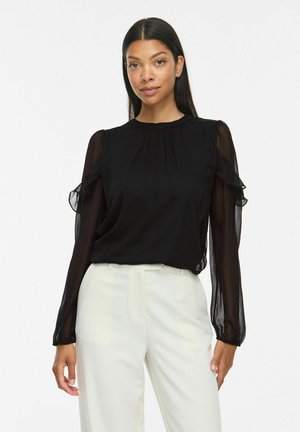 Femme aux longs cheveux foncés porte un chemisier noir transparent à manches longues avec des volants et un pantalon blanc taille haute ajusté sur fond blanc.