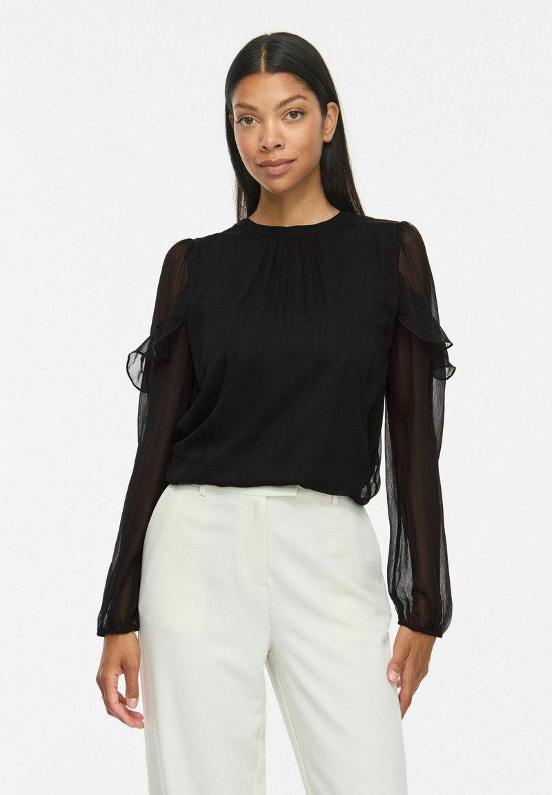 Femme aux longs cheveux foncés porte un chemisier noir transparent à manches longues avec des volants et un pantalon blanc taille haute ajusté sur fond blanc.
