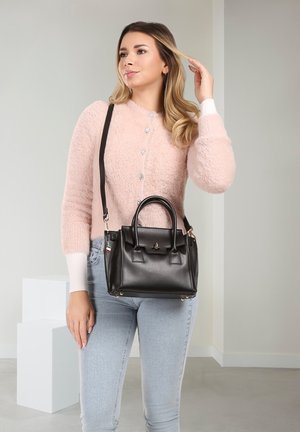 Woman wearing pink fuzzy sweater and light jeans, holding black leather handbag with shoulder strap in minimalist indoor setting.