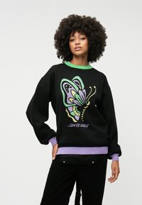 Sweater preto com um design colorido de borboleta em verde, roxo e amarelo, com punhos e barra em verde e roxo canelados. O texto diz "SANTA CRUZ."
