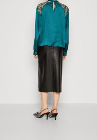 Blouse en satin sarcelle avec des détails en dentelle sur les épaules, dos à ouverture en trou de serrure, associée à une jupe en cuir noire longueur genou et des talons à motifs.