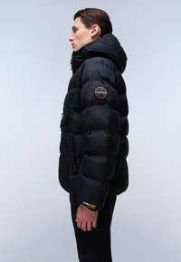 Giacca a vento nera con cappuccio, con finitura lucida, un'apertura rotonda sul braccio superiore e un'etichetta 'CORDURA®' sulla manica.