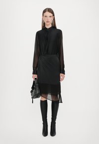Black sheer blouse with a button closure, paired with a black skirt featuring a layered transparent hem. Knee-high black boots and a studded handbag.