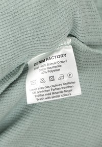 Vaateetiketti vaaleansinisellä tekstuurilla, jossa näkyy materiaalikoostumus ja monikieliset pesuohjeet huoltokuvakein kanssa.