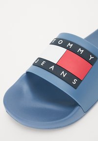 Kék csúszó szandál széles pánttal, fekete, fehér és piros blokkban megjelenő logográfival, textúrált felülettel és sima talppal.