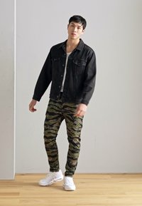 Jachetă din denim negru peste un tricou alb, asortată cu pantaloni kaki cu model de camuflaj și sneakers albi. Texturi netede, croi casual și modele distincte.