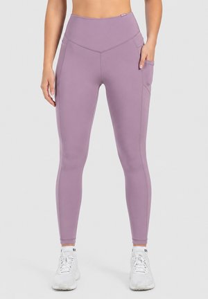 Femme portant des leggings lavande taille haute avec poches latérales et des baskets blanches, debout devant un fond clair uni.
