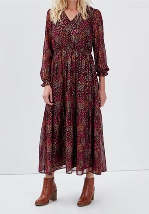 Mujer con un vestido burdeos floral de manga larga y largo hasta el tobillo, combinado con botines marrones de tacón sobre un fondo liso.