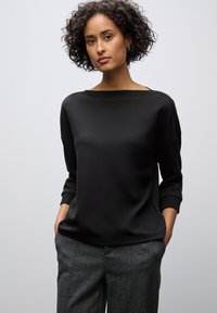 Haut noir à manches longues avec un décolleté large, associé à un pantalon gris texturé et à motifs. La tenue a une coupe décontractée et un design minimaliste.