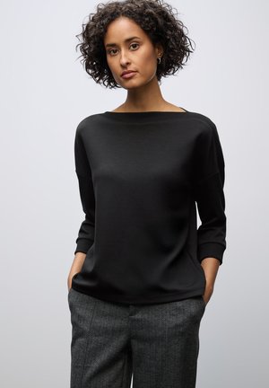 Haut noir à manches longues avec un décolleté large, associé à un pantalon gris texturé et à motifs. La tenue a une coupe décontractée et un design minimaliste.
