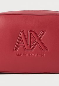 Raudona dirbtinės odos piniginė su iškilusiu "AX" logotipu ir "Armani Exchange" tekstu, pasižyminti lygia tekstūra ir stačiakampiu pavidalu.
