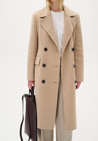 Gaiši beidžu vilnas mētelis ar dubulto pogu aizdari, melnām pogām un sānu kabatām, valkāts virs baltas T-kreklu un gaišām biksēm, apvienots ar tumšu somu.