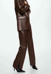Osoba w ciemno-brązowym skórzanym garniturze z długim żakietem i szerokimi spodniami, w połączeniu z czarnymi szpilkami w kształcie migdałów.