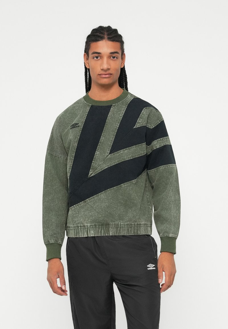 Fiatal férfi, fonott hajjal, zöld és fekete mintás pulóverben és fekete Umbro nadrágban, egy fehér háttér előtt áll.