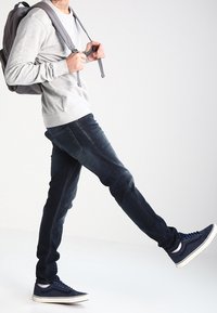 Sudadera gris claro, vaqueros ajustados azul oscuro y zapatillas azul marino. El modelo lleva una mochila gris y camina sobre un fondo neutro.