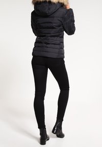 Czarna puchowa kurtka z kapturem wykończonym sztucznym futrem, pikowanym wzorem i zapięciem na zamek. Noszona na czarnych obcisłych dżinsach i botkach za kostkę.