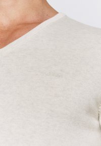 Beige V-Ausschnitt T-Shirt aus geripptem Baumwollstoff. Verfügt über dezente Logo-Stickerei und eine glatte Textur. Keine sichtbaren Muster oder Akzente.
