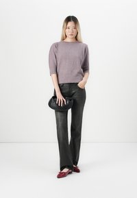 Ung kvinne står mot hvit bakgrunn iført lilla genser, svarte bukser, røde sko, holder en svart clutch-veske med den ene hånden i lommen.