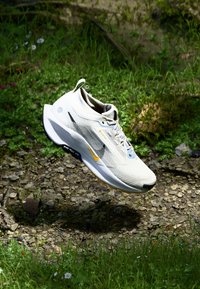 Chaussure de course légère en blanc et gris, dotée d'une tige en maille texturée, d'accents bleus, d'une semelle noire et d'un logo jaune sur le côté.
