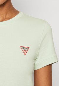 Světl zelené bavlněné tričko s červeným a bílým trojúhelníkovým logem na levé straně hrudi. Krátké rukávy a kulatý výstřih.