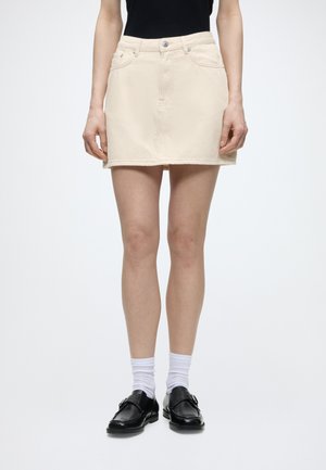 Femme portant une mini-jupe en jean beige, un haut noir, des chaussettes blanches et des mocassins en cuir noirs, debout devant un fond uni.