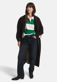 Brun frakk, grønn og hvit stripepolo, mørke jeans og brune støvler. Frakken har en løs passform og går til kneet.