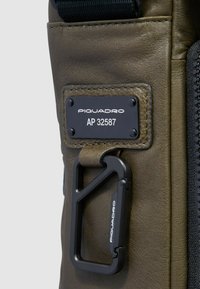 Bolsa de cuero verde oliva con una placa metálica negra que lleva grabado el nombre de la marca y el número de modelo, junto a un mosquetón negro para su sujeción.