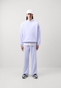 Lila oversized pulóver hímzett logóval, hozzáillő lila nadrággal párosítva. A megjelenést egy fehér sapka és mintás fekete-fehér cipő egészíti ki.