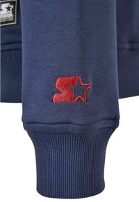 Tengerészkék pulóver anyag ribbed mandzsettákkal; piros hímzett logóval, amely egy csillagot és az 'S' betűt ábrázolja. Simább tapintású, klasszikus szabású.