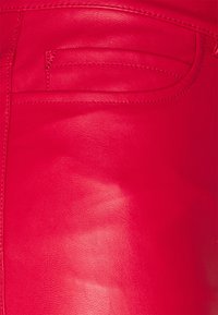 Rode neplederen broek met een gladde textuur, voorzien van een voorzak met subtiele stikdetails. Close-up weergave van het stofoppervlak.