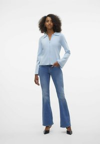 Svetlo modra zip-up jakna s ovratnikom in zvonastimi rokavi, kombinirana z razširjenimi modrimi kavbojkami in črnimi špičastimi čevlji. Enostaven dizajn z gladko teksturo.