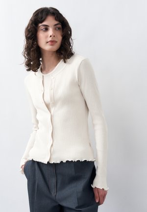 Młoda kobieta o kręconych włosach, ubrana w białą prążkowaną kardigan i ciemne spodnie z wysokim stanem, stojąca na tle jednolitego jasnego tła.