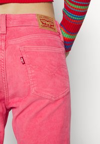 Personne portant un jean en velours côtelé rose Levi's avec poche arrière et écusson de marque marron, associé à un haut rayé à manches longues multicolore.