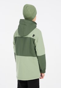 Giacca verde con cappuccio, caratterizzata da un design a blocchi di colore in verde chiaro e scuro. Berretto a maglia a costine nello stesso colore.
