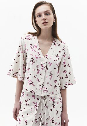 Jeune femme portant un haut ample blanc et un pantalon avec un motif floral rose et bordeaux, debout devant un fond blanc.