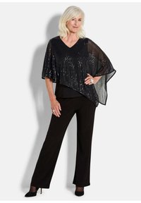 Schwarzer Pailletten-Poncho mit V-Ausschnitt, über einem schwarzen Oberteil und weit geschnittenen Hosen getragen. Mit transparenter Textur und asymmetrischem Schnitt.